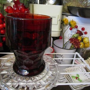 May include: A ruby red glass tumbler with a faceted base sits on a clear, decorative glass plate. The tumbler is partially filled and has a wide, open top. The background includes other kitchen items.