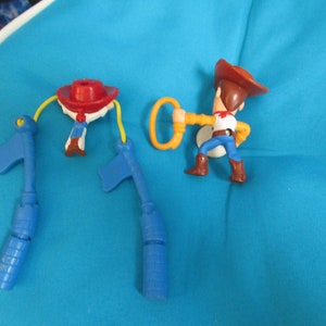 May include: A set of three blue plastic toy figures, including Woody from Toy Story, with a yellow lasso and a red hat. The figures are on a blue surface.