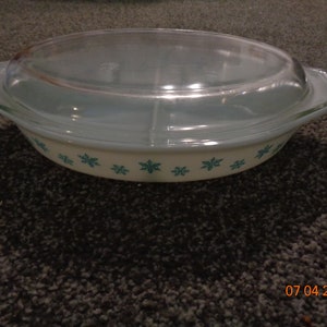 May include: An oval, vintage Pyrex casserole dish with a clear glass lid. The dish is off-white with teal snowflake designs around the base. The lid and handles are teal. A classic piece for baking and serving.