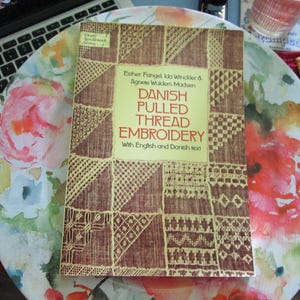 May include: A book titled "Danish Pulled Thread Embroidery" with English and Danish text. The cover features a brown and beige geometric pattern. The book is on a floral patterned surface, with a laptop keyboard visible in the background.
