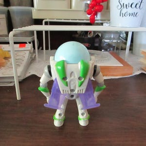 K&ouml;nnte beinhalten: Eine wei&szlig;e und gr&uuml;ne Buzz Lightyear Actionfigur mit einem hellblauen Helm und lila Fl&uuml;geln. Die Figur steht auf einer braunen Oberfl&auml;che.