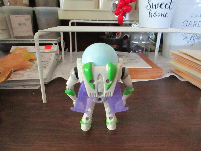 K&ouml;nnte beinhalten: Eine wei&szlig;e und gr&uuml;ne Buzz Lightyear Actionfigur mit einem hellblauen Helm und lila Fl&uuml;geln. Die Figur steht auf einer braunen Oberfl&auml;che.