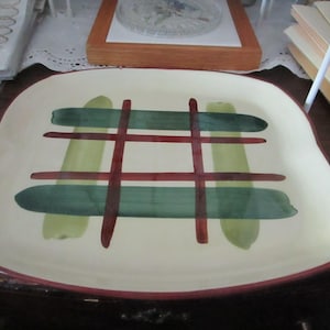 May include: A white ceramic serving platter with a red, green, and brown plaid design. The platter has a slightly rounded shape and a red rim.
