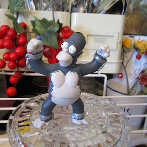 May include: A figurine of a cartoon character in a gorilla costume, with a gray body, white chest, and white hands and feet. The figure is standing on a clear, patterned glass surface. The background includes red berries and containers labeled "Thyme" and "Parsley."