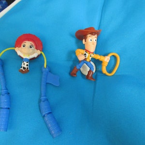 May include: Two plastic toy figures of Woody and Jessie from the Toy Story franchise. Woody is wearing a brown hat and a yellow shirt. Jessie is wearing a red hat and a blue and white shirt. Both figures are on a blue background.