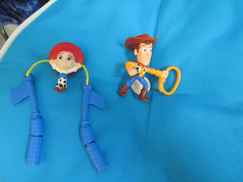 May include: Two plastic toy figures of Woody and Jessie from the Toy Story franchise. Woody is wearing a brown hat and a yellow shirt. Jessie is wearing a red hat and a blue and white shirt. Both figures are on a blue background.