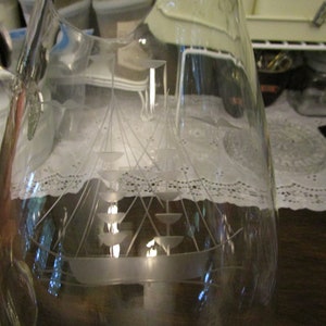 May include: A clear glass pitcher with an etched design of a sailboat. The pitcher has a handle and a wide mouth.
