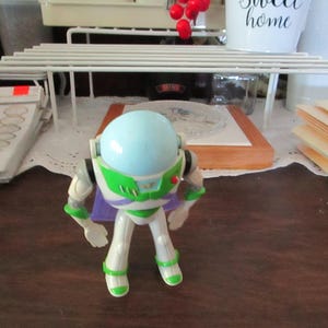 K&ouml;nnte beinhalten: Eine wei&szlig;e und gr&uuml;ne Buzz Lightyear Actionfigur mit einem hellblauen Helm und einem lila Umhang. Die Figur steht auf einer Holzoberfl&auml;che.