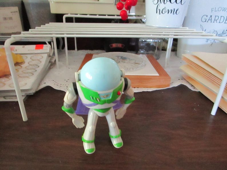 K&ouml;nnte beinhalten: Eine wei&szlig;e und gr&uuml;ne Buzz Lightyear Actionfigur mit einem hellblauen Helm und einem lila Umhang. Die Figur steht auf einer Holzoberfl&auml;che.