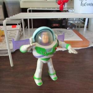 K&ouml;nnte beinhalten: Eine Plastik-Spielzeugfigur von Buzz Lightyear aus der Toy Story-Reihe. Die Figur tr&auml;gt einen wei&szlig;en und gr&uuml;nen Raumanzug mit einem violetten Visier und einem blauen Helm. Die Figur steht mit ausgestreckten Armen.