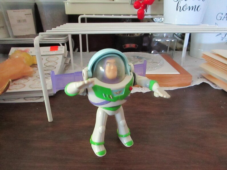 K&ouml;nnte beinhalten: Eine Plastik-Spielzeugfigur von Buzz Lightyear aus der Toy Story-Reihe. Die Figur tr&auml;gt einen wei&szlig;en und gr&uuml;nen Raumanzug mit einem violetten Visier und einem blauen Helm. Die Figur steht mit ausgestreckten Armen.