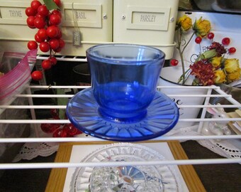 May include: A cobalt blue glass teacup and saucer set. The cup has a rounded shape and sits on a matching saucer with a ridged design. The set is displayed on a white wire shelf, with red berries and dried flowers in the background.