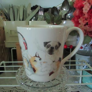May include: White ceramic mug featuring a pug and a cat illustration. The mug has a curved handle and sits on a clear glass coaster. The background includes silverware, flowers, and other decorative items.