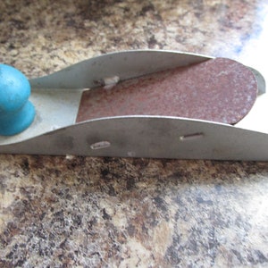 May include: A small, metal hand plane with a blue wooden handle. The plane has a rusty metal blade and is used for smoothing wood.