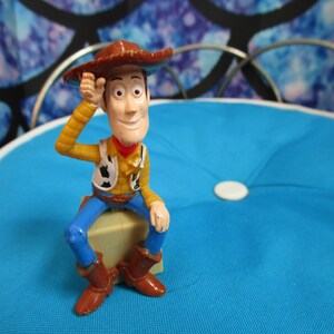 Pode incluir: Uma figura de a&ccedil;&atilde;o do Woody, vestida com chap&eacute;u de cowboy, camisa amarela, colete, cal&ccedil;as azuis e botas castanhas, senta-se num bloco de cor clara. A figura posa com uma m&atilde;o no chap&eacute;u. O fundo &eacute; uma almofada azul.