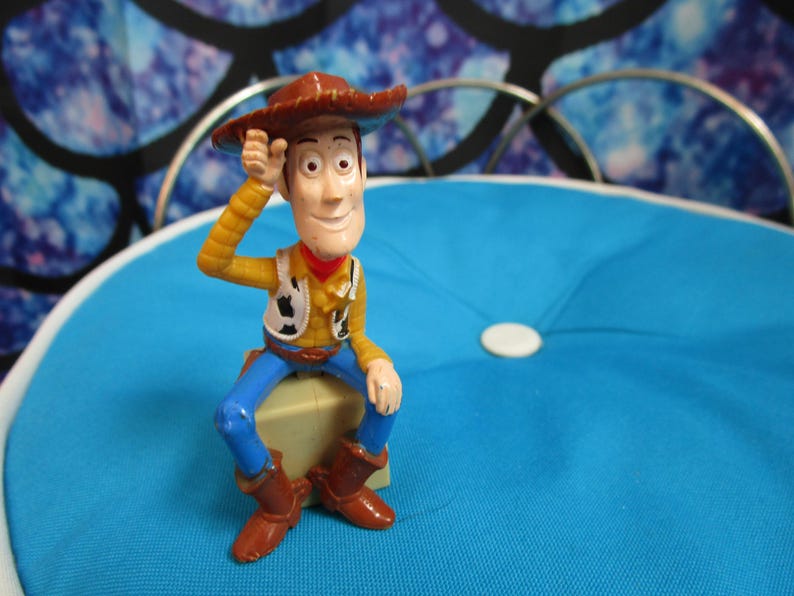 Pode incluir: Uma figura de a&ccedil;&atilde;o do Woody, vestida com chap&eacute;u de cowboy, camisa amarela, colete, cal&ccedil;as azuis e botas castanhas, senta-se num bloco de cor clara. A figura posa com uma m&atilde;o no chap&eacute;u. O fundo &eacute; uma almofada azul.