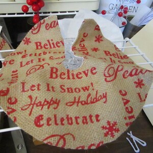 May include: A round burlap Christmas tree skirt with red text that reads "Believe", "Let It Snow", "Happy Holiday", and "Celebrate". The skirt has a hole in the center for the tree trunk and snowflake designs.