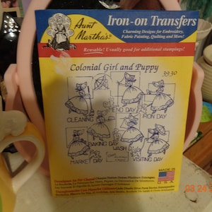 May include: Iron-on transfer sheet with a design of a colonial girl and puppy. The sheet is yellow with blue and black text. The design is for embroidery, fabric painting, quilting, and more. The sheet is reusable and is made in the USA.