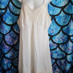 May include: A white slip dress with lace trim around the neckline and hem. The dress has thin straps and a v-neck.