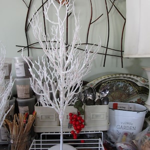 May include: A white twig tree with white glitter on the branches. The tree is on a white stand and is surrounded by various kitchen items, including jars, a white metal container with the word "Parsley" on it, and a white metal container with the words "Flower Garden" on it.