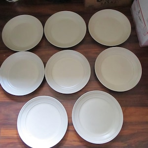 May include: Nine off-white ceramic plates arranged on a dark wood surface. Each plate has a slightly raised rim and a smooth, matte finish. The plates are evenly spaced, suggesting a set for dining or serving.
