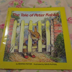 May include: A yellow book cover with the title "The Tale of Peter Rabbit" in black text. The cover features an illustration of Peter Rabbit peeking through a wooden fence. The book is by Beatrix Potter and illustrated by David McPhail.