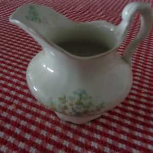 May include: A white ceramic creamer with a floral design and a scalloped edge. The creamer has a handle and is sitting on a red and white checkered tablecloth.