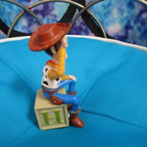Pode incluir: Uma figura do personagem Woody da franquia Toy Story. Woody usa um chap&eacute;u de cowboy marrom, camisa amarela, colete branco com manchas pretas, cal&ccedil;as azuis e botas marrons. Ele est&aacute; sentado em um bloco bege com a letra "H".