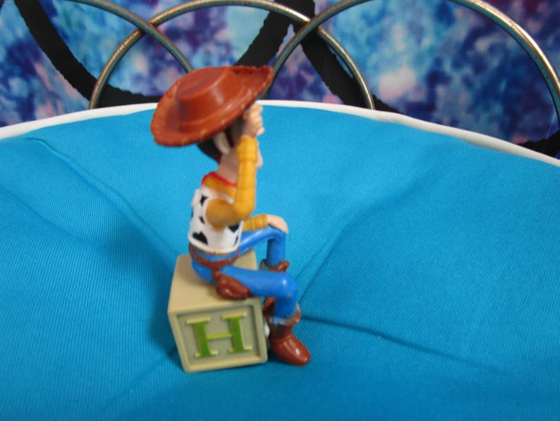 Pode incluir: Uma figura do personagem Woody da franquia Toy Story. Woody usa um chap&eacute;u de cowboy marrom, camisa amarela, colete branco com manchas pretas, cal&ccedil;as azuis e botas marrons. Ele est&aacute; sentado em um bloco bege com a letra "H".