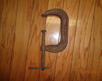 May include: A rusty metal C-clamp with a threaded screw and a handle. The clamp is on a wooden surface.