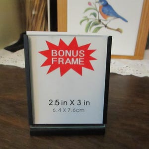 May include: A black picture frame with a white insert. The insert has a red starburst with the words "BONUS FRAME" and the dimensions 2.5 in x 3 in, and 6.4 x 7.6cm. The frame is on a wooden surface.