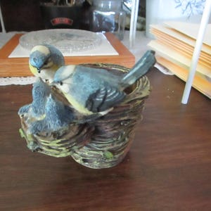 May include: A bluebird figurine perched on a branch with a nest. The bird is painted with blue, white, and yellow accents. The nest is made of brown and green material.