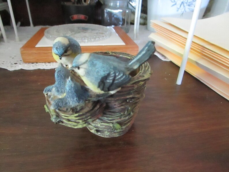 May include: A bluebird figurine perched on a branch with a nest. The bird is painted with blue, white, and yellow accents. The nest is made of brown and green material.
