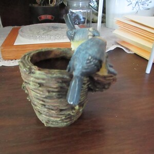 May include: A small, brown, woven planter with two blue birds perched on the rim. The birds are painted with white and yellow accents.