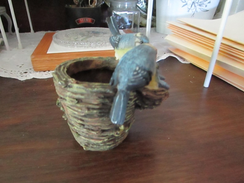 May include: A small, brown, woven planter with two blue birds perched on the rim. The birds are painted with white and yellow accents.