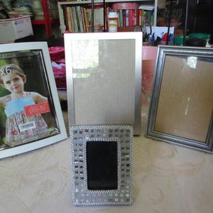 May include: Four picture frames of varying styles and sizes. One frame is white with a 4x6 photo, another is silver, and a third is a mosaic design. The frames are empty, ready for photos.