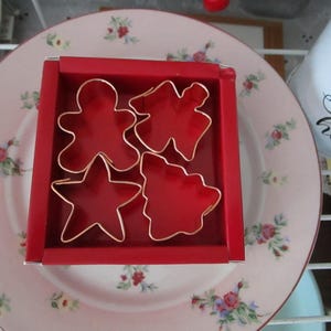 May include: A red box with four cookie cutters inside. The cutters are shaped like a gingerbread man, a star, a Christmas tree, and an angel. The box is sitting on a pink plate with a floral pattern.