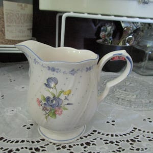 May include: A white ceramic creamer with a blue and floral design. The creamer has a scalloped edge and a handle.