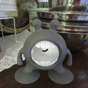 May include: A gray, cartoon-like, monster-shaped, silicone suction cup holder with a large, white, toothy mouth. The holder has two suction cups on the bottom.