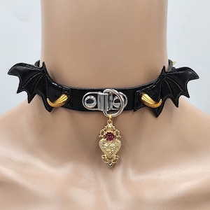 Puede incluir: Gargantilla de cuero negro con alas de murciélago doradas y un colgante de corazón dorado con una joya roja.