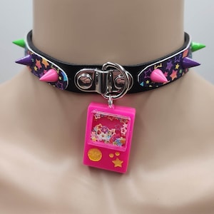Pu&ograve; includere: Un collare choker nero con dettagli argentati e una stampa di stelle colorate. Il choker ha punte verdi e rosa. Un ciondolo rosa con una finestra trasparente piena di stelle colorate &egrave; appeso al collare.