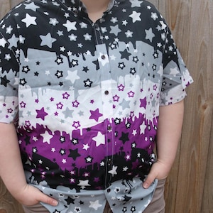 May include: A short-sleeved button-up shirt with a black, gray, white, and purple print of stars and dripping paint. The shirt has a collar and a button-up front.