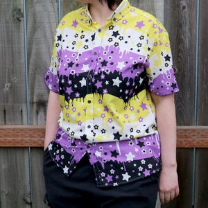 Könnte beinhalten: Ein kurzärmeliges Hemd mit Knöpfen und einem farbenfrohen Druck aus Sternen und Streifen in Gelb, Lila und Schwarz. Das Hemd hat eine lockere Passform und einen Kragen.