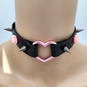 Puede incluir: Collar gargantilla negro con un centro en forma de corazón rosa y púas plateadas. El collar tiene pequeños detalles de rosas rosas. El collar está hecho de cuero negro.