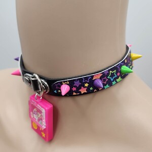 Pu&ograve; includere: Un collare choker nero con stelle arcobaleno e punte colorate. Un ciondolo rosa con una finestra trasparente &egrave; attaccato al collare.