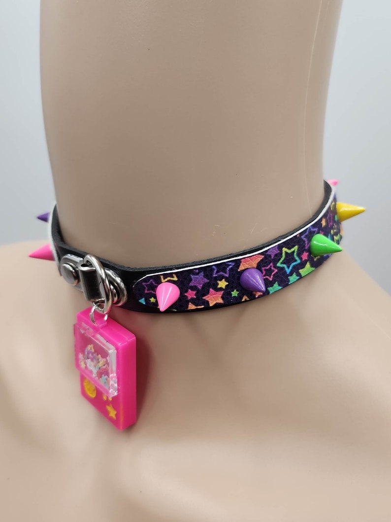 Pu&ograve; includere: Un collare choker nero con stelle arcobaleno e punte colorate. Un ciondolo rosa con una finestra trasparente &egrave; attaccato al collare.