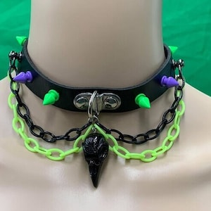 May include: A black leather choker with green and purple spikes, a black chain, and a lime green chain. A black crow skull pendant hangs from the choker.