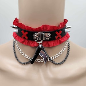 Puede incluir: Un collar gargantilla rojo y negro con herrajes plateados, pinchos blancos y un dije plateado. La gargantilla tiene un borde de volantes rojos y dos X rojas en la correa de cuero negro.