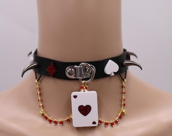 May include: A black leather choker necklace with silver spikes and a red glitter heart playing card charm. The necklace also features a gold chain with red beads.