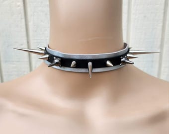 May include: A layered choker necklace featuring a black band centred over a silver band. The choker is adorned with multiple metallic silver-toned spikes of varying lengths, including two long spikes extending from the sides and shorter spikes along the front.